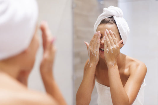 A woman with a towel wrapped around her head is washing her face with a gentle cleanser in front of a bathroom mirror.