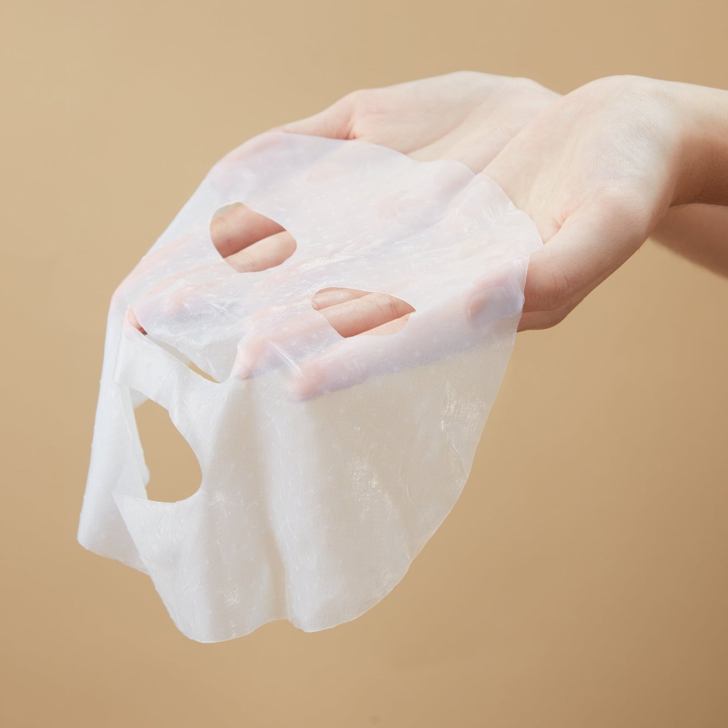 Soft, translucent facial sheet mask held in hands against a neutral background.