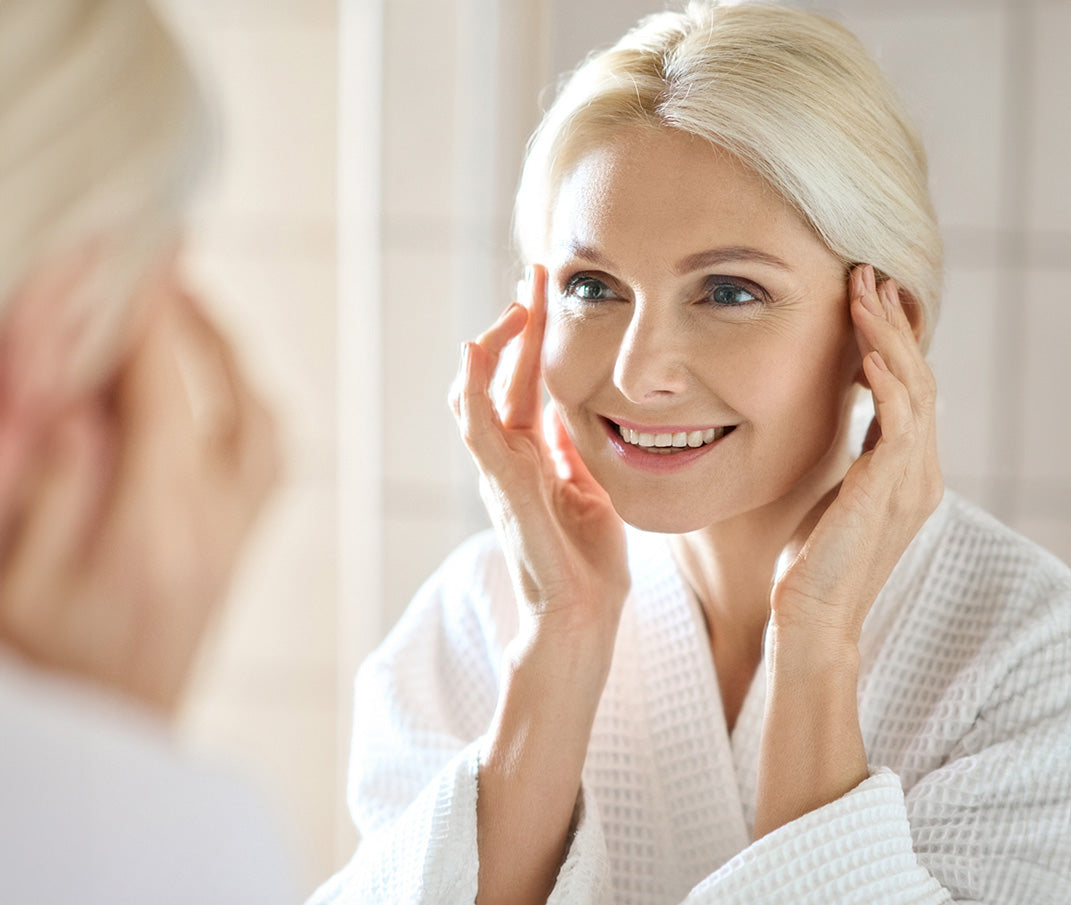 Smiling woman in bathrobe gently touching her face, admiring her reflection in the mirror.