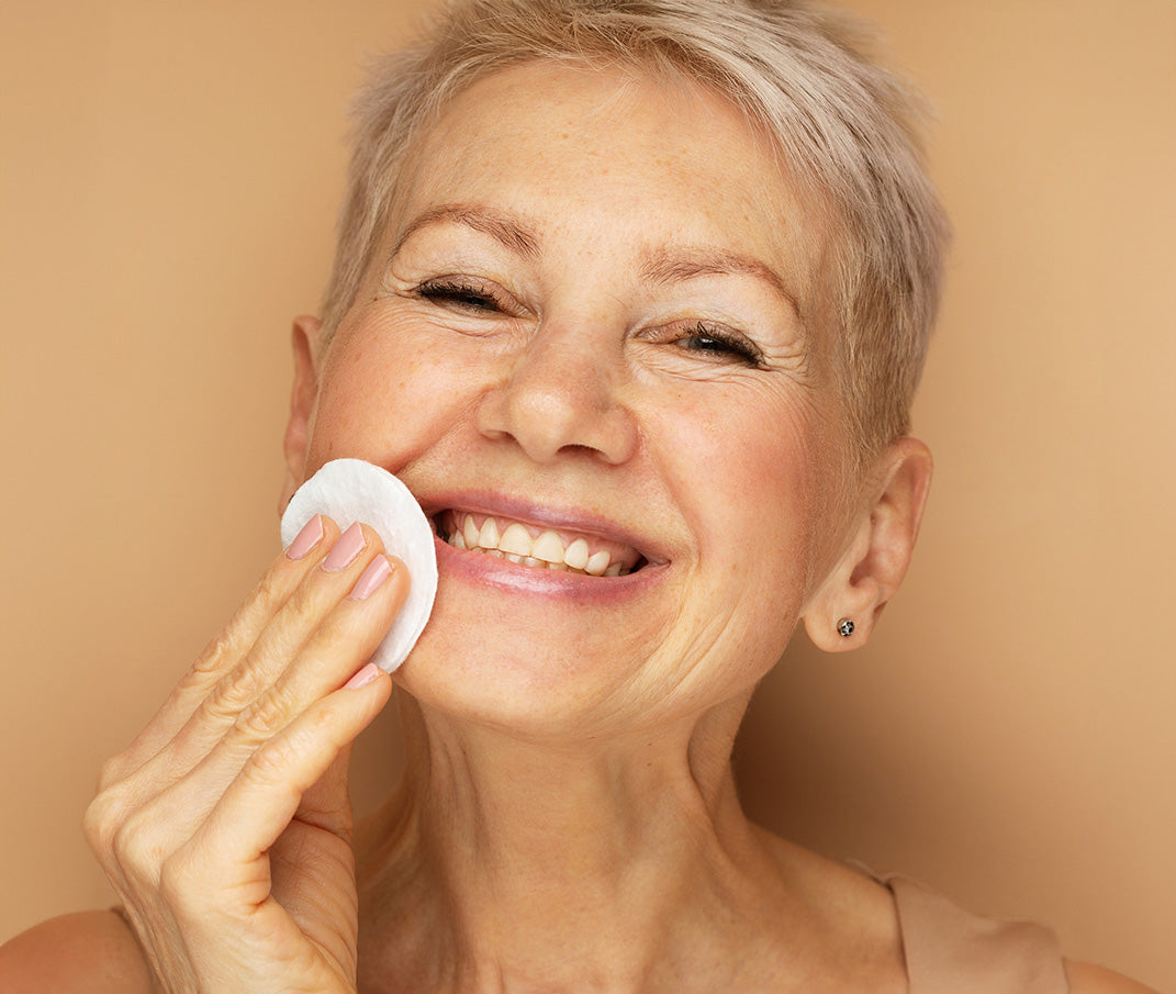 Smiling mature woman applying skincare with cotton pad.