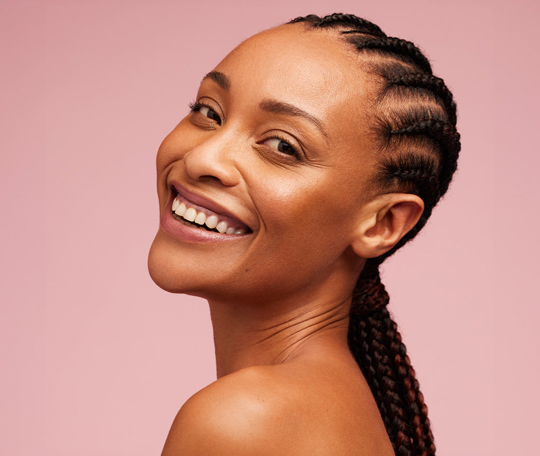 Woman with radiant skin and cornrows smiling against a pink background.