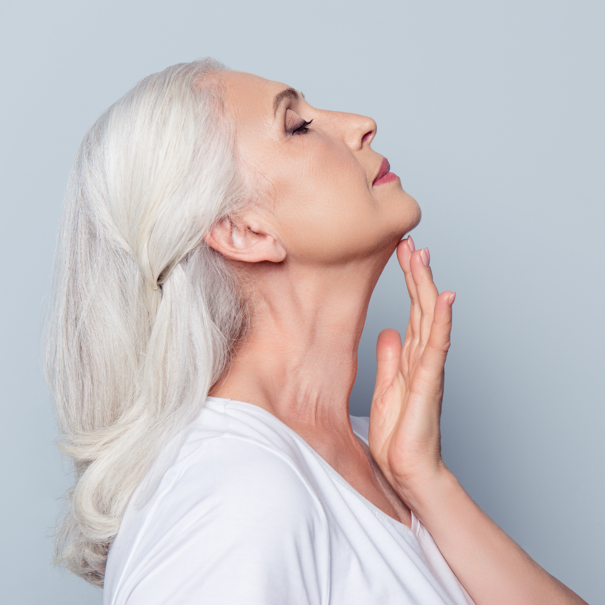 Close-up profile of a woman gently touching her neck and chin, showcasing healthy, firm skin against a light gray background.