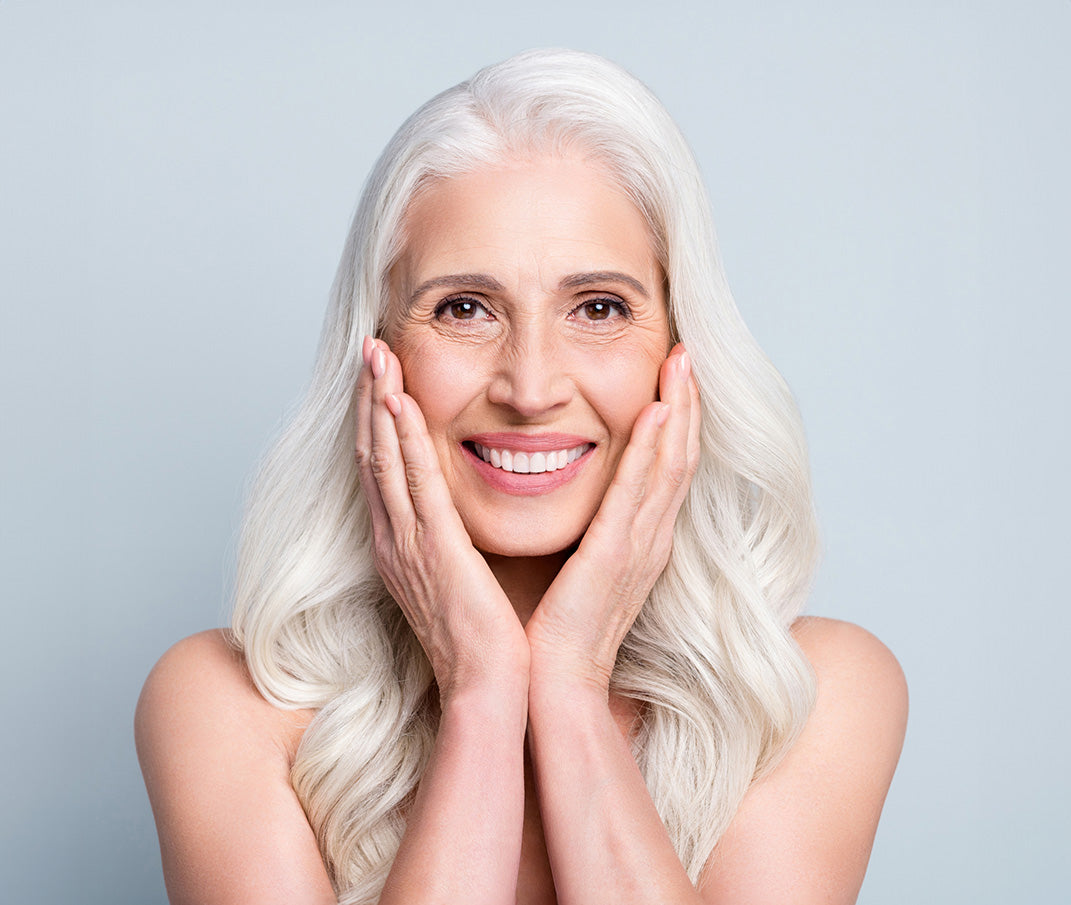 Smiling woman with long gray hair, showcasing healthy skin against a light blue backdrop.