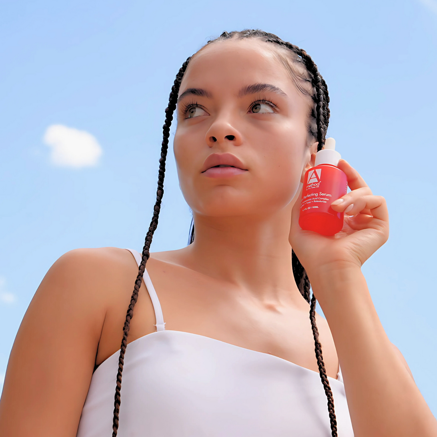 Woman with braids holding a red perfecting serum bottle against a bright blue sky.