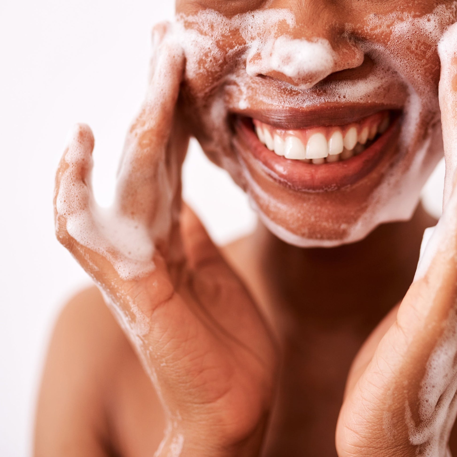 Close-up of a person's face covered in rich, cleansing foam, smiling during a skincare routine.