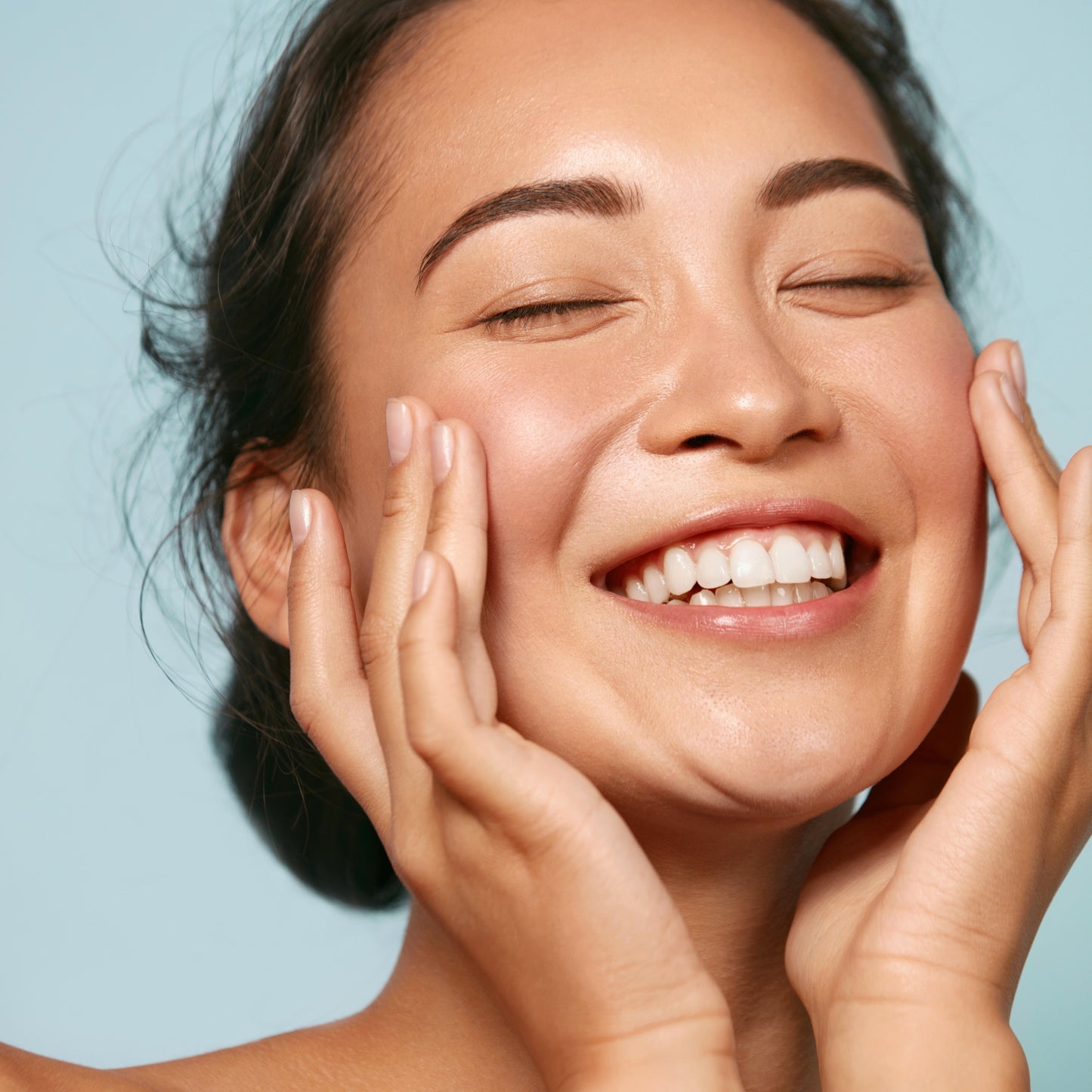 Close-up of a smiling woman gently touching her face, showcasing healthy, radiant skin against a light blue backdrop.