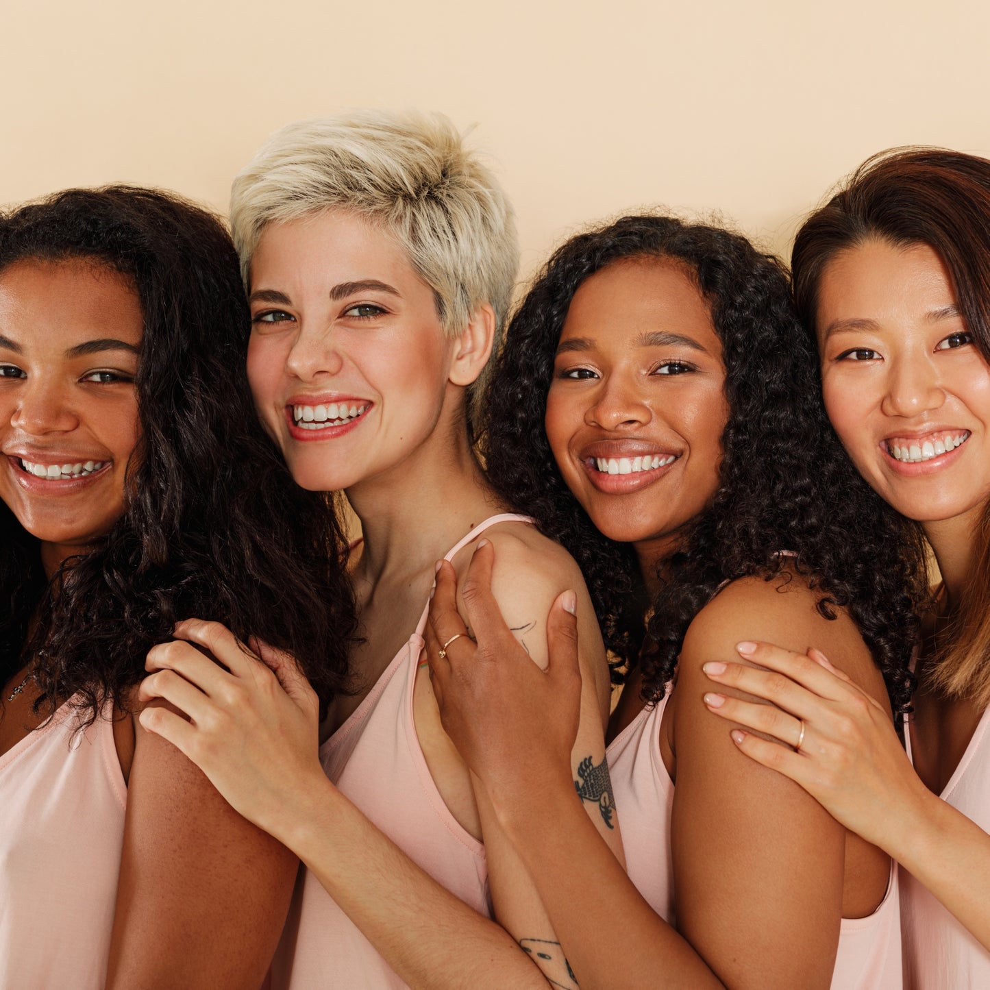 Diverse group of women with varying skin tones, smiling and embracing against a beige background.