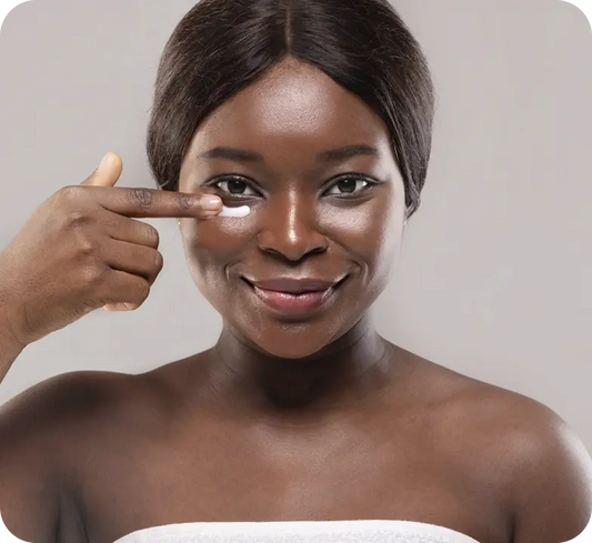 A woman with dark skin applies a white cream from her finger to her under-eye area. She is wearing a white towel wrapped around her shoulders against a solid light gray background.