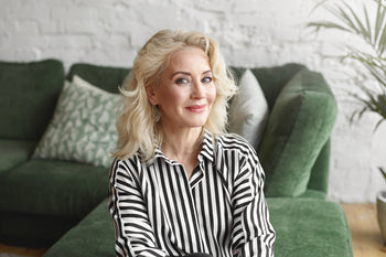 Dermatologist-recommended skincare routine illustrated by a confident middle-aged woman with healthy, radiant skin sitting in a bright, modern living space.