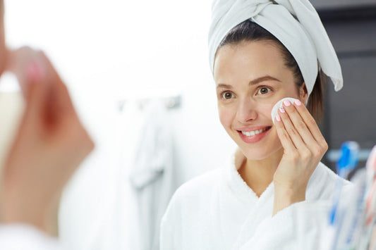Woman in a white bathrobe and towel gently using a cotton pad on her face during a double cleanse skincare routine in front of a bathroom mirror.