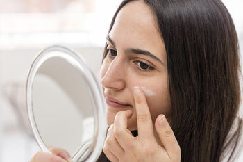 Woman applying blemish control cream to her cheek while looking in a mirror.