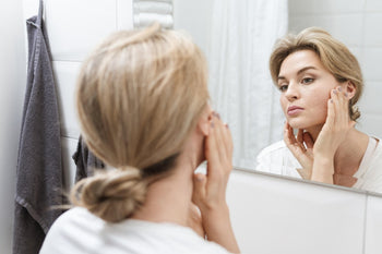 Woman examining the skin on her face in a bathroom mirror.