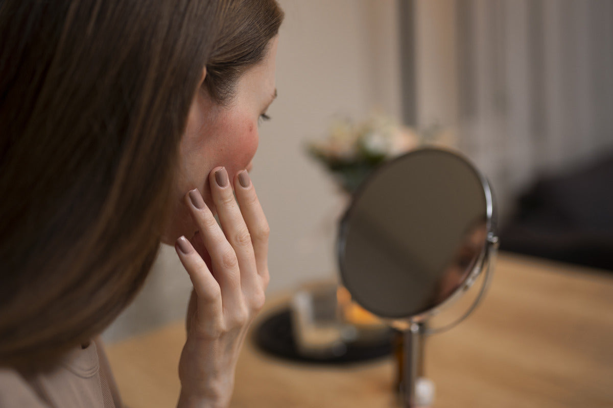 Woman examining facial skin in a mirror while touching her cheek. Image concept for barrier repair cream.