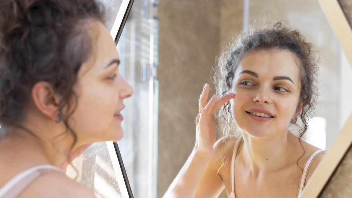 Woman with curly hair smiling in the mirror while applying skincare cream to her face.