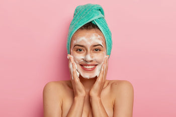 Smiling woman with a towel wrapped around her hair gently massaging a foaming cleanser for combination skin onto her face against a pink background.