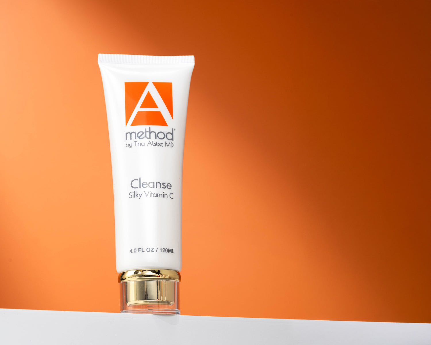The A Method Silky Vitamin C Cleanser displayed on a white platform with an orange backdrop for holiday gifting.